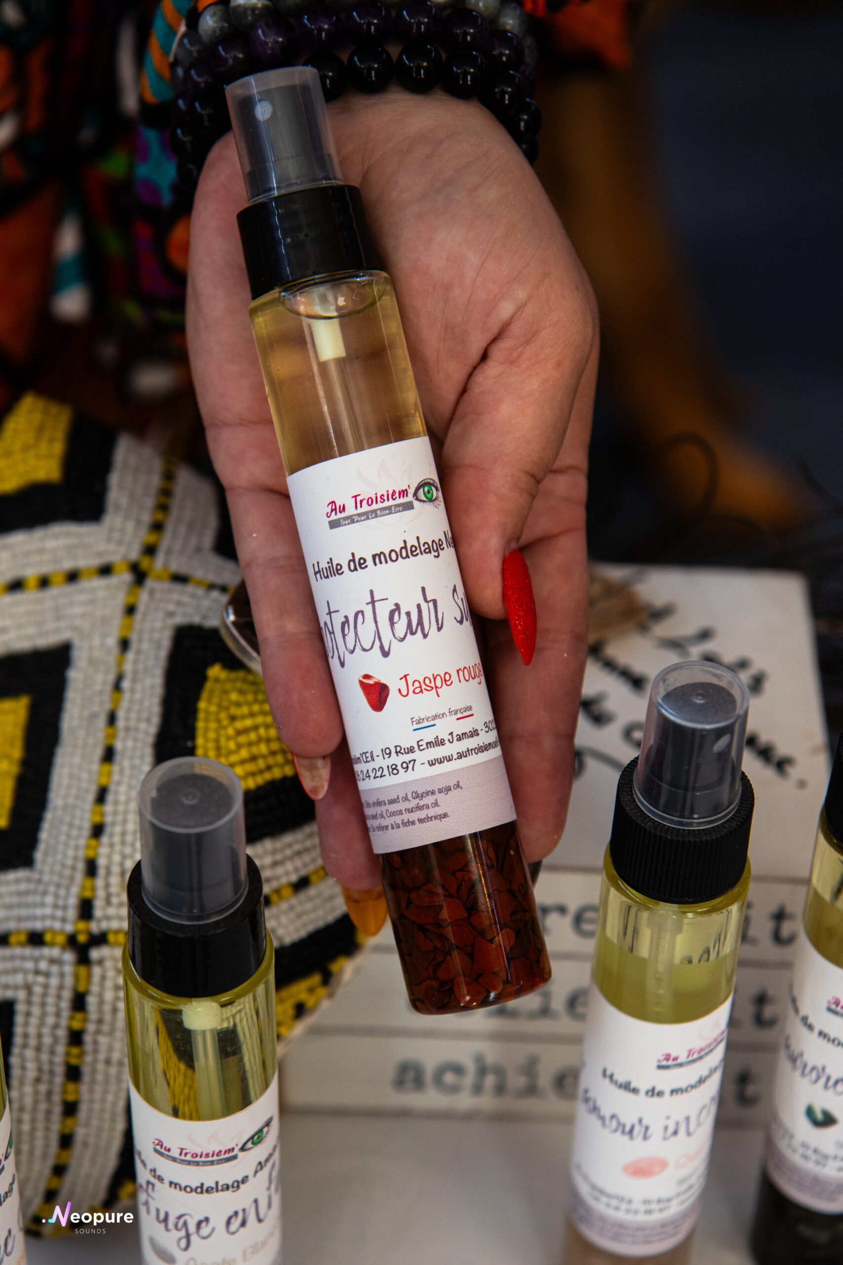 Flacon spray d’huile de modelage avec étiquettes “Au Troisien” et contenu rouge, tenu par une main, produits bien visibles. Flacon spray d’huile de modelage avec étiquettes “Au Troisien” et contenu rouge, tenu par une main, produits bien visibles.